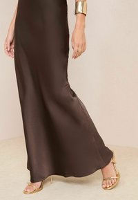 Donkerbruine satijnen maxi-jurk met een gladde textuur, gecombineerd met metallic gouden sandalen met bandjes en een hoge hak.