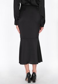 Zwarte midi-rok met een aansluitende pasvorm, uitlopende zoom en een gladde textuur. Draagt zwarte hoge hakken. Achteraanzicht weergegeven.