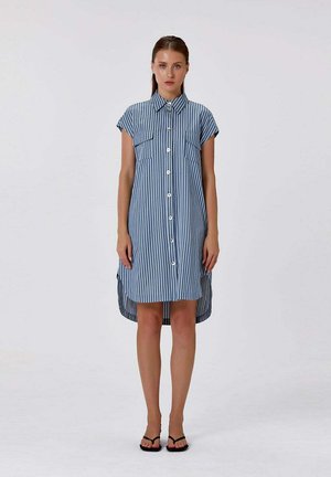Femme debout portant une robe chemise à rayures verticales bleues et blanches à manches courtes avec des boutons à l'avant et des sandales noires sur un fond uni.