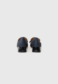 Chaussures bleu marine avec un motif de pois texturé, présentant un bout arrondi et un petit talon noir brillant. Vue arrière affichée.