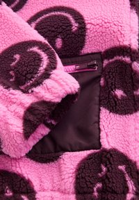 Tissu polaire rose et marron foncé avec motif de visages souriants et une poche zippée violette.