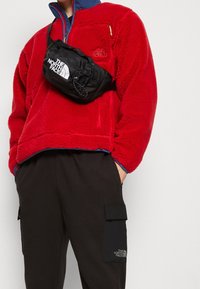 Sudadera de forro polar roja con cuello azul, que cuenta con un bolsillo con cremallera. Riñonera negra con logo, llevada sobre pantalones cargo negros con bolsillos laterales.