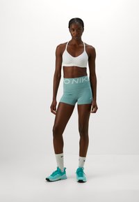 Lyseblå Nike shorts med hvit midjebånd, hvit sports-BH og turkise treningssko. Modellen står mot en enkel hvit bakgrunn.