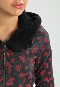 Zwarte hoodiejack met een zachte, fleece-gevoerde kraag en een grijze body met rode bladranden. Ritssluiting met decoratieve hardware.