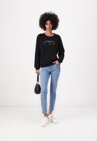 Sudadera negra con cuello redondo y diseño gráfico. Combinada con jeans de color azul claro y zapatillas blancas. El modelo sostiene un bolso negro pequeño.