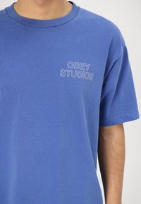 Modra bombažna majica s kratkimi rokavi in okroglim izrezom. Na levi strani prsnega dela ima belo vezeno besedilo "OBEY STUDIOS".