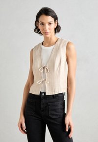 Beige gestreepte crop top met een V-hals en een strikdetail aan de voorkant, gecombineerd met zwarte high-waisted jeans. Sofa textuur, mouwloos ontwerp.