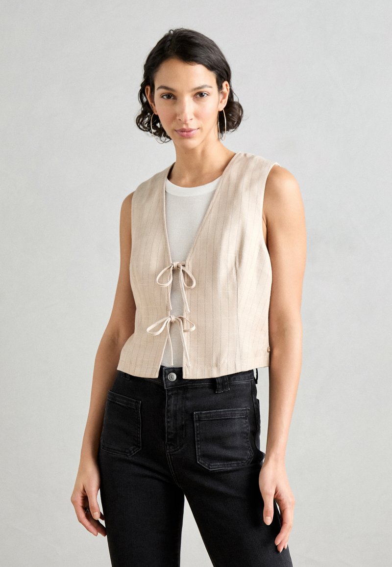 Beige gestreepte crop top met een V-hals en een strikdetail aan de voorkant, gecombineerd met zwarte high-waisted jeans. Sofa textuur, mouwloos ontwerp.