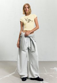 Žluté zkrácené tričko s černou grafikou klokanů, spárované s volnými šedými tepláky a šedým svetr, který je uvázaný v pase. Černá obuv.