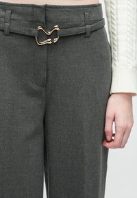 Nahaufnahme von grauen Hosen mit einer herzförmigen goldenen Gürtelschnalle und dem Ärmel eines weißen Strickpullovers am rechten Arm einer Person.