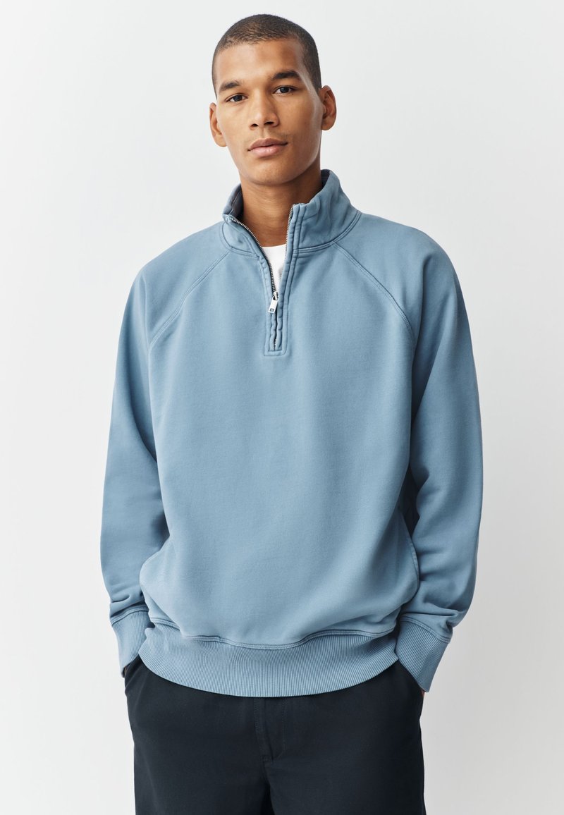 Noormehe lühikeste juustega, kes kannab helesinist neljandiku tõmblukuga pusa ja musti pükse, seisab tavalise valge tausta ees.