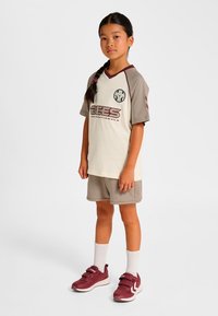 Jugendsportoutfit in hellbeige und braun mit einem "BEES"-Logo, bestehend aus einem V-Ausschnitt-Shirt, passenden Shorts und kastanienbraunen Sneakers mit Klettverschlüssen.