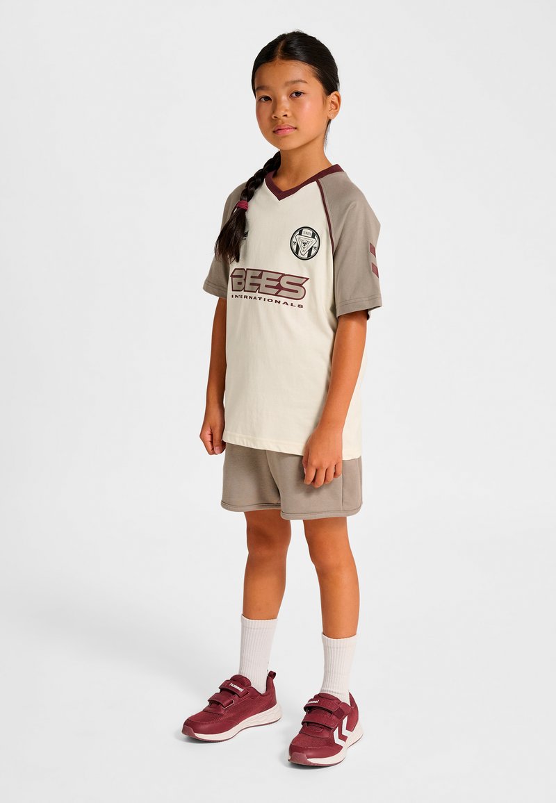 Jugendsportoutfit in hellbeige und braun mit einem "BEES"-Logo, bestehend aus einem V-Ausschnitt-Shirt, passenden Shorts und kastanienbraunen Sneakers mit Klettverschlüssen.