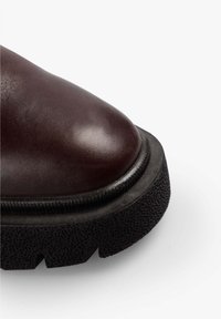 Bota de cuero marrón oscuro con punta redondeada, suela de goma negra texturizada y detalle de borde en contraste alrededor de la suela.