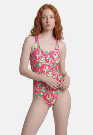 Young woman with red hair wearing a colorful green and pink patterned one-piece swimsuit with double pink shoulder straps.