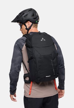 Man draagt een zwarte fietshelm en zonnebril en draagt een zwarte Vaude Trail Control 18L rugzak met borst- en taillebanden.