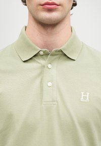 Polo verde claro com um tecido texturizado, três botões no colarinho e um pequeno logótipo branco "H" no lado esquerdo do peito.
