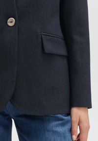 Blazer marine avec une texture lisse, fermeture à bouton unique et une poche latérale. Associé à un jean en denim, présentant une coupe ajustée.