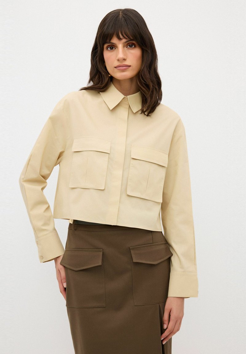 Camicia beige tagliata con colletto a punta e due tasche sul petto; abbinata a una gonna marrone con tasche a patta e uno spacco laterale.