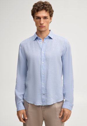 Mann trägt ein hellblaues Langarm-Hemd und beige Hose, steht vor einem schlichten hellen Hintergrund.