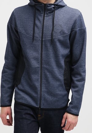 Uomo che indossa una felpa con cappuccio blu navy con pannelli laterali neri e coulisse, abbinata a jeans scuri, in piedi davanti a uno sfondo neutro.