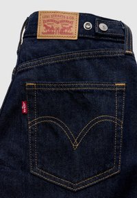 Σκούρα μπλε τζιν τζιν με δερμάτινη ετικέτα με ανάγλυφο το "Levi Strauss & Co.". Χρυσοκέντητη ραφή περιγράφει τις πίσω τσέπες με μια κόκκινη ταμπέλα.