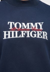 Μπλε ναυτικό φούτερ με ψηλό γιακά, με κεντημένο το κείμενο "TOMMY HILFIGER" σε άσπρο και κόκκινο χρώμα. Μαλακή υφή και άνετη γραμμή.