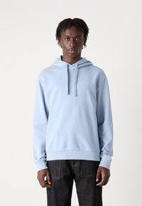 Sudadera azul claro con cordón, puños y dobladillo acanalados. Fabricada con una tela suave, presentada en un modelo con jeans oscuros y cabello texturizado.