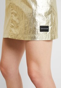 Gonna in laminato oro con una texture stropicciata, caratterizzata da un'etichetta nera Calvin Klein Jeans sull'orlo. Design corto e aderente.