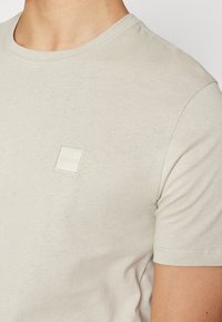 Gros plan d'une personne portant un T-shirt beige clair avec un petit patch logo carré indiquant "BOSS" sur la poitrine.