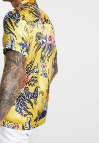 Koszula z krótkim rękawem w żółtym kolorze z wzorem kwiatów i liści w czerni, różu i pomarańczy. Gładka tekstura z luźnym krojem.