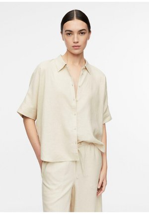 Woman with dark hair tied back wearing a loose beige short-sleeve button-up shirt and matching drawstring pants against a plain white background.