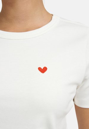 Weißes T-Shirt mit Rundhalsausschnitt und einem kleinen roten, gestickten Herz auf der linken Brust, getragen von einer Person.