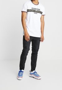 Valkoinen lyhythihainen paita, jossa on musta "GLORIOUS" -teksti ja kreikkalainen avainkuvio, yhdistettynä kuluneisiin mustiin farkkuihin ja monivärisiin lenkkareihin.