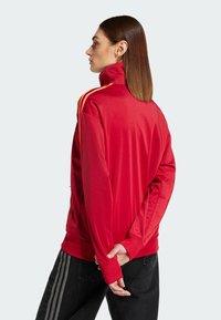 Rote Sportjacke mit hohem Kragen, mit orangen Streifen an den Ärmeln, aus glattem Material gefertigt, mit lockerer Passform und Reißverschlusstaschen.
