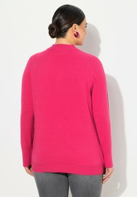 Fuksia neule, jossa on pitkät hihat ja pyöreä kaulus. Neulottu pinta, jossa on ribbireuna. Yhdistetty harmaisiin housuihin, nähtynä takaa.