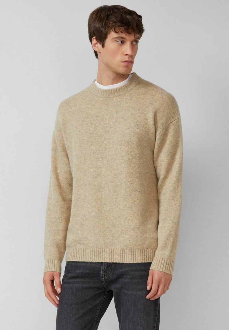 Beige gebreide trui met een ronde hals, geribde manchetten en zoom, gemaakt van zachte stof. Draagt met een donkere spijkerbroek, tegen een neutrale achtergrond.