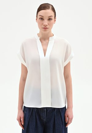 Jeune femme aux cheveux lissés vers l'arrière, portant un chemisier blanc à manches courtes avec un décolleté en V profond et un pantalon plissé foncé, debout devant un fond uni.