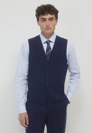 Jeune homme aux cheveux bouclés portant un gilet à rayures navy, un pantalon assorti, une chemise rayée bleu clair et une cravate à rayures navy, se tenant contre un mur clair.