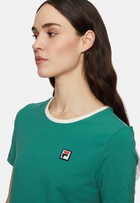 T-shirt in cotone verde con maniche corte, colletto bianco e logo ricamato in rosso, bianco e blu sul petto. Design semplice.