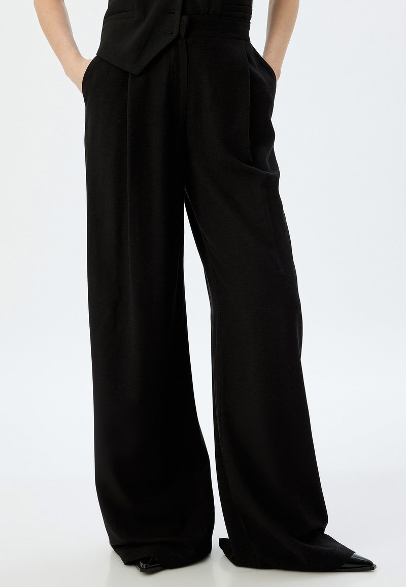Schwarze weit geschnittene Hose aus glattem Stoff mit hoher Taille, seitlichen Taschen und einem lässigen Schnitt, der am Knöchel schmaler zuläuft.