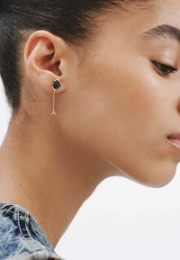 Pendientes de gota de piedra verde con un acabado metálico en tono dorado; presentan una piedra cuadrada y una cadena con un acento triangular.