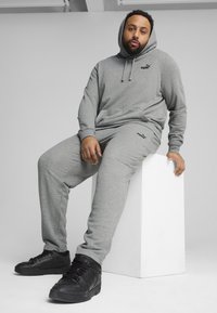 Grauer Sweatshirt und Jogginghose aus weichem Baumwollmix, mit Kapuze und seitlichen Taschen. Schwarze Logo-Details auf beiden Teilen. Darunter schwarze Sneaker.