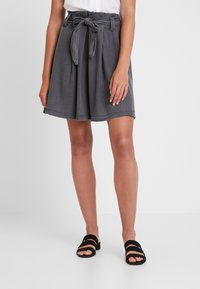 Femme portant un short plissé taille haute gris foncé avec une ceinture nouée, un haut blanc et des sandales noires ouvertes à deux brides.
