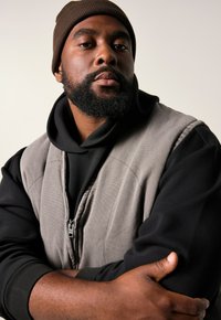 Graue ärmellose Weste über einem schwarzen Kapuzenpullover, mit einer gerippten braunen Mütze. Nahaufnahme des Gesichts mit verschränkten Armen, die strukturierte Materialien zeigen.