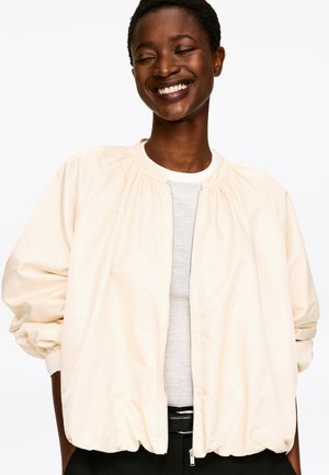 Veste bomber beige clair avec épaules froncées, fermeture éclair à l'avant et poignets élastiques. Portée sur un haut gris clair, avec une coupe décontractée.