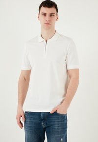 Vit polo-shirt med dragkedja i kragen, korta ärmar och texturerade accenter, i kombination med blå jeans. Slitstekt tyg, minimalistisk design.