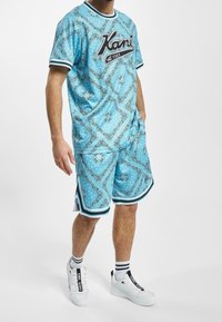 Hombre que lleva una camisa de manga corta azul con un patrón y pantalones cortos a juego, calcetines blancos con rayas negras y zapatillas blancas con detalles en negro.