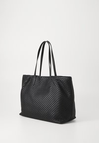 Sac fourre-tout noir tissé avec doubles anses d'épaule, base rectangulaire et bordure lisse, posé contre un fond clair uni.