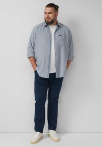 Jasnoniebieska koszula na długi rękaw z guzikami i kieszenią, noszona na białym t-shircie, zestawiona z granatowymi spodniami i białymi butami sportowymi.